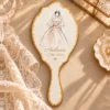 Oglindă de mână albă cu margine aurie, cu schiță de modă a unei mirese în rochie Oglindă de mână albă cu margine aurie, cu schiță de modă a unei mirese în rochie cloș cu roz-somon și voal scurt, numele în auriu